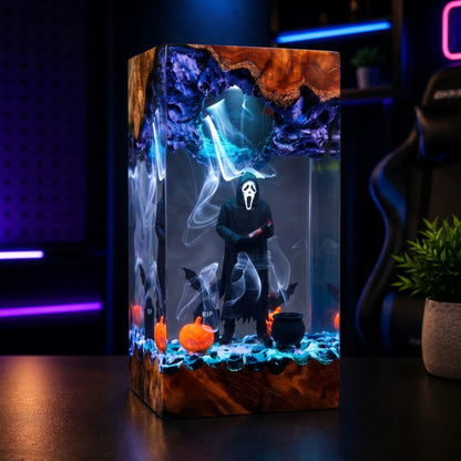 A rectangular LED light box featuring a Ghostface Halloween design, displayed on a dark table in a gaming room setting. The box is illuminated with blue and purple lights, showing a figure in a black robe with a white mask, surrounded by spooky graphics like bats, a cauldron, and orange pumpkins.